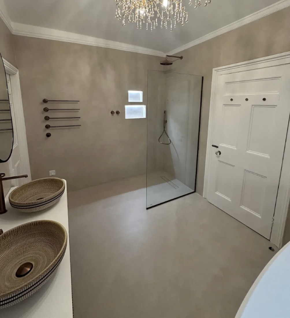 Microcement wet room with a walk-in glass shower, twin woven vessel basins and a crystal chandelier